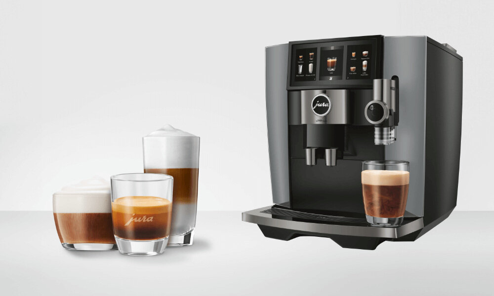Na obrazie widoczny jest ekspres do kawy z logo \'Jura\' oraz trzy szklanki z różnymi rodzajami kawy, w tym cappuccino i espresso. Ekspres ma nowoczesny design z wyświetlaczem i dyszą do spieniania mleka.