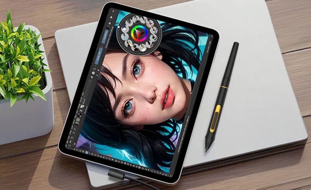 Na stole leży tablet graficzny z wyświetlonym kolorowym portretem kobiety, obok znajduje się rysik. W tle widoczna jest doniczka z zieloną rośliną.