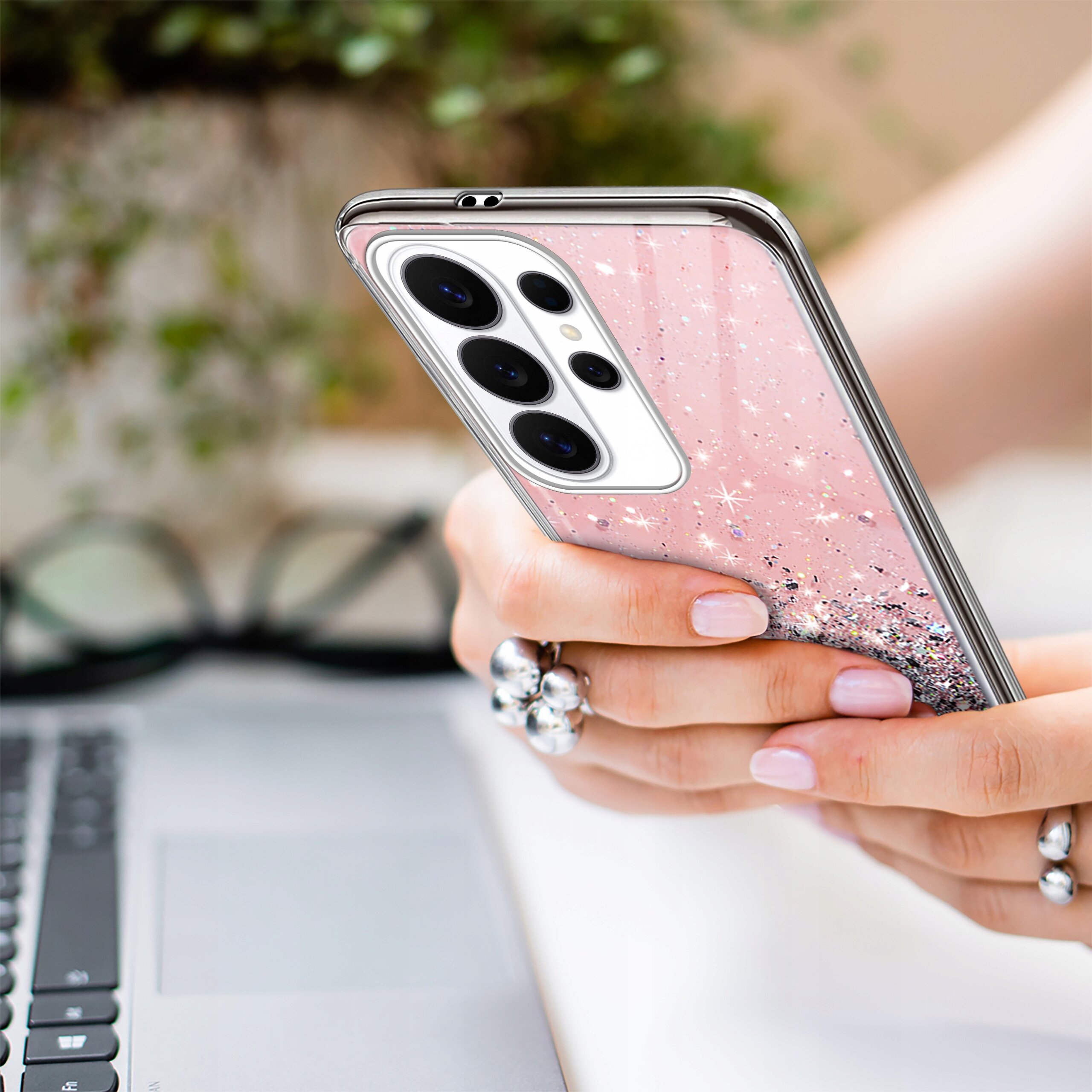 Osoba trzyma telefon w różowym etui z brokatem, na tle klawiatury laptopa i okularów. Etui ma przezroczyste krawędzie i widoczne trzy obiektywy aparatu.