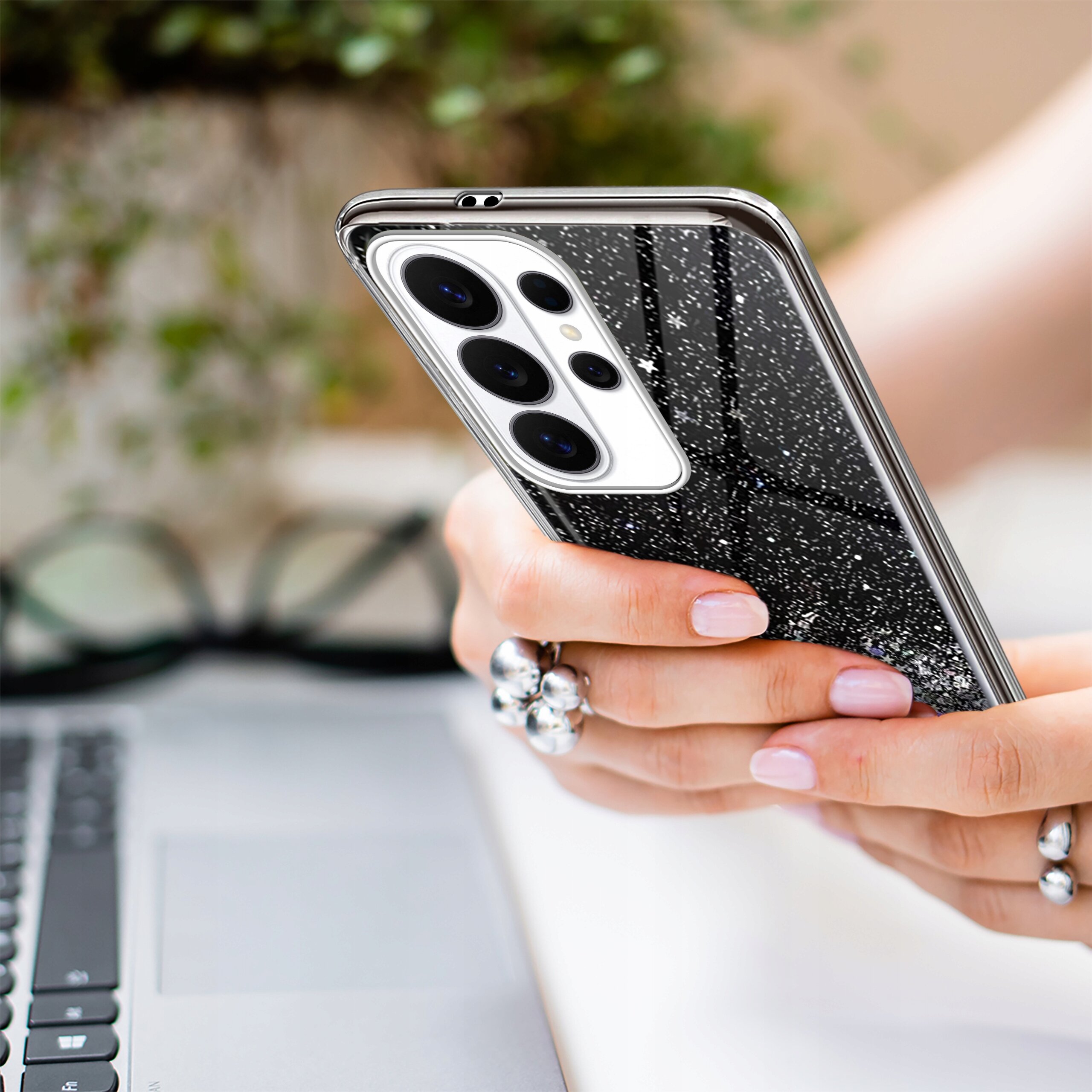 Osoba trzyma telefon w etui z brokatowym wzorem, obok znajduje się klawiatura laptopa. W tle widoczne są rozmyte elementy dekoracyjne.