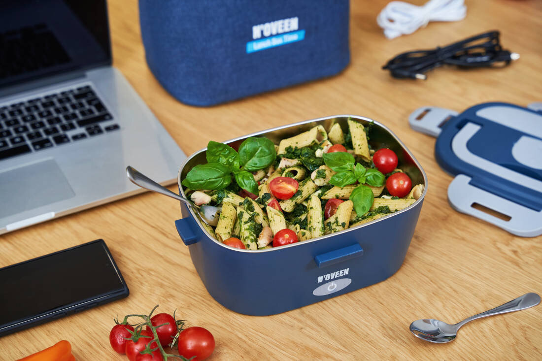 Na drewnianym stole znajduje się elektryczny lunch box marki NOVEEN wypełniony makaronem z zielonym sosem, pomidorkami koktajlowymi i liśćmi bazylii. Obok leży pokrywka, łyżka, laptop, smartfon oraz torba z napisem \'NOVEEN Electric Lunch Box\'.
