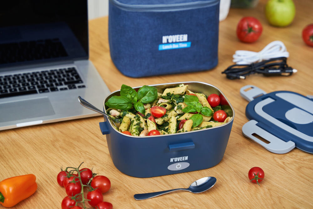 Na drewnianym stole znajduje się elektryczny lunch box NOVEEN wypełniony jedzeniem, obok leży łyżka i pokrywka. W tle widoczna jest torba z napisem \'NOVEEN Lunch Box Time\', laptop oraz kilka warzyw.