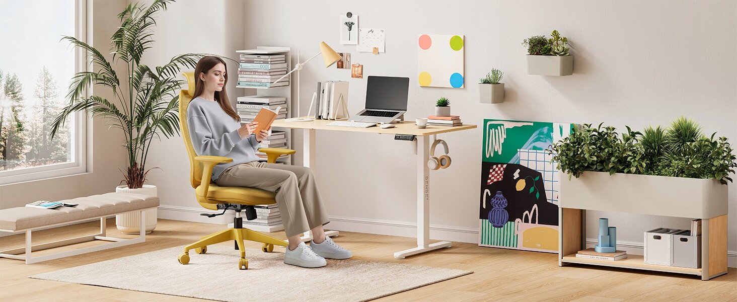 Osoba siedzi na żółtym, ergonomicznym krześle biurowym przy biurku w nowoczesnym, jasnym wnętrzu z roślinami i dekoracjami na ścianie. Na biurku znajduje się laptop, książki i inne akcesoria biurowe.