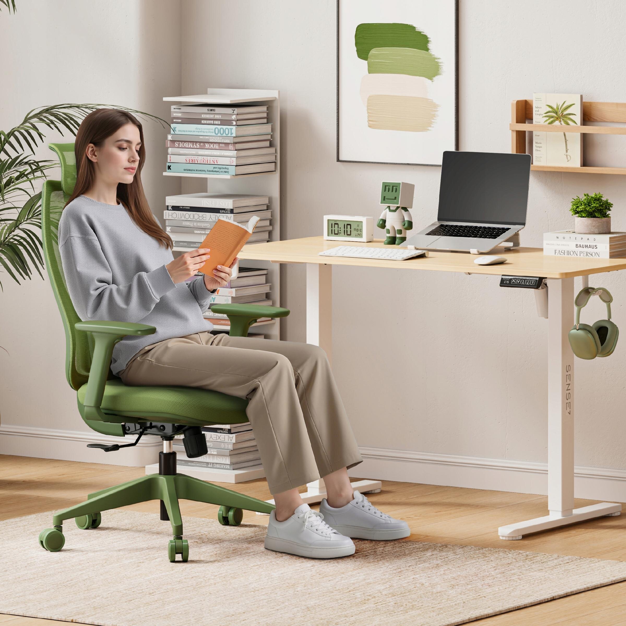 Osoba siedzi na zielonym, ergonomicznym fotelu biurowym przy biurku z laptopem i zegarem. W tle widać półkę z książkami i rośliną oraz obraz z kolorowymi paskami.