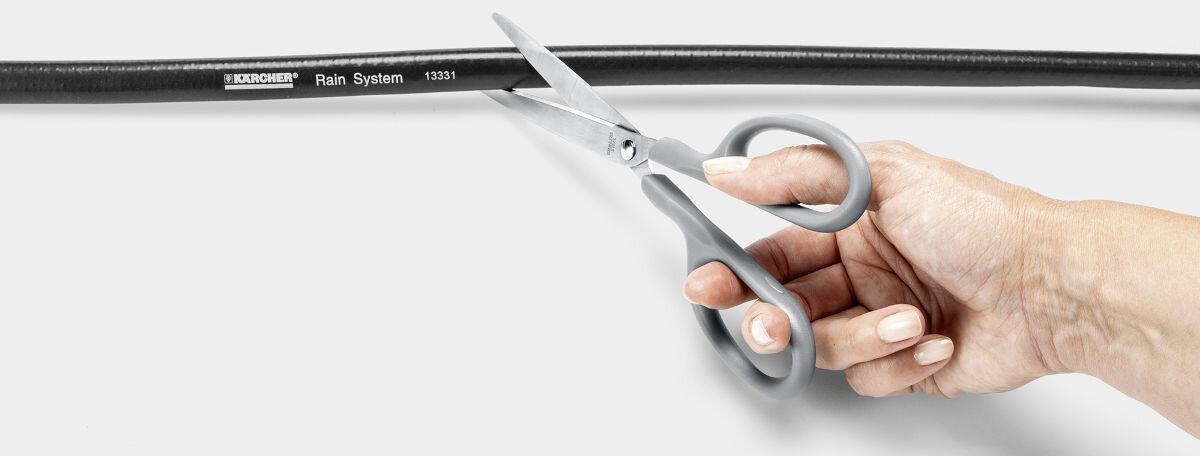 Zestaw do nawadniania KARCHER Rain System 2.645-238.0, elementy, rozbudowanie, aranżacja, ogród, na białym tle dłoń trzyma nożyczki i przycina wąż do potrzebnych wymiarów