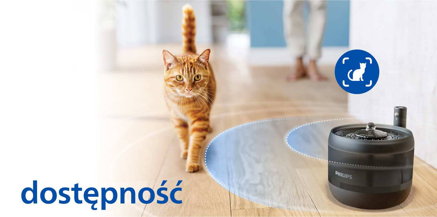 Na drewnianej podłodze stoi czarna fontanna dla zwierząt z logo PHILIPS, obok której znajduje się kot. W lewym dolnym rogu widoczny jest napis \'dostępność\'.