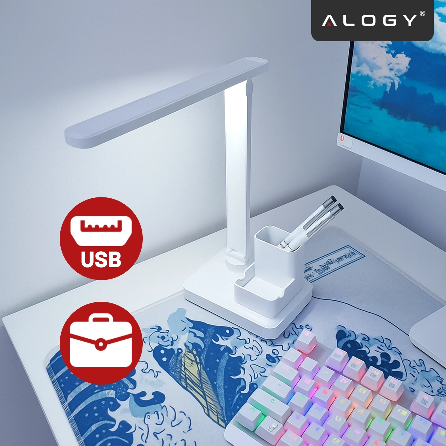 Na biurku znajduje się biała lampka biurkowa LED z organizerem, obok której leży klawiatura z podświetleniem RGB. W tle widoczny jest monitor z obrazem nieba i chmur, a w rogu zdjęcia znajduje się logo \'ALOGY\'.