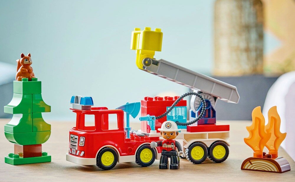 Zestaw LEGO przedstawia czerwony wóz strażacki z wysięgnikiem, figurkę strażaka, drzewo z wiewiórką oraz płomienie. W tle widoczne są elementy dekoracyjne wnętrza.