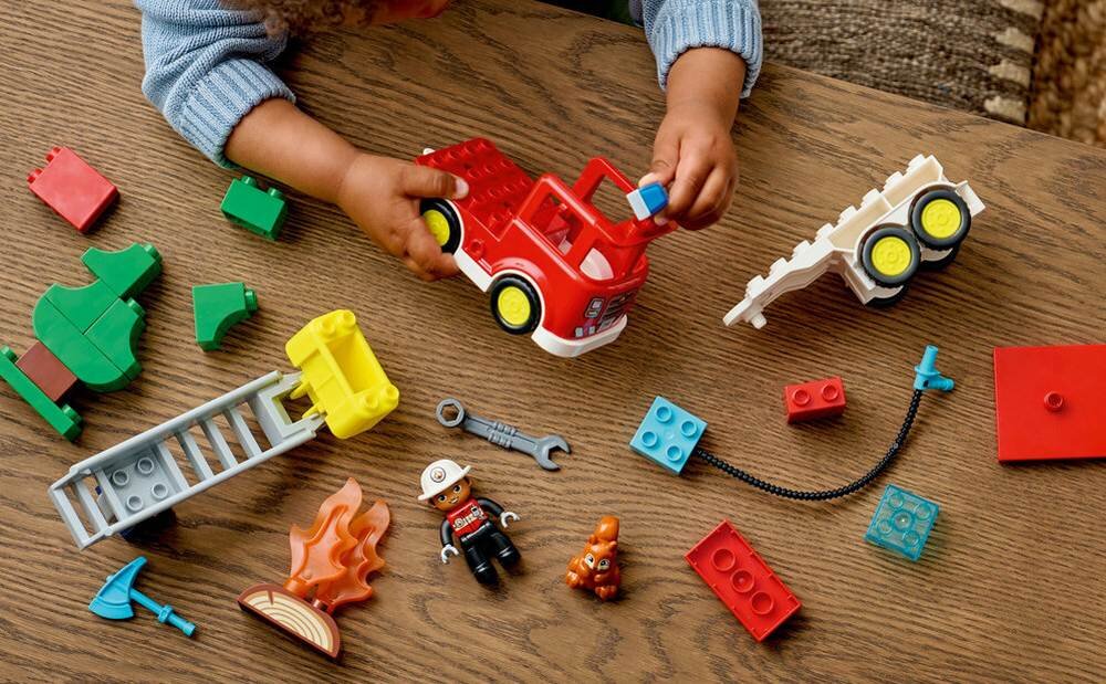Na drewnianym stole znajdują się klocki LEGO, w tym czerwony wóz strażacki, figurka strażaka, drabina i elementy płomieni. Dziecko bawi się klockami, trzymając wóz strażacki.
