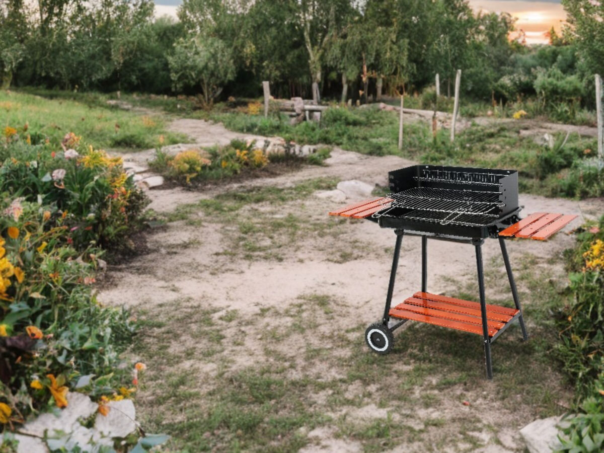 Grill węglowy na kółkach z półkami bocznymi i regulacją wysokości rusztu stoi na trawiastej ścieżce w ogrodzie. W tle widoczne są drzewa i kwiaty.