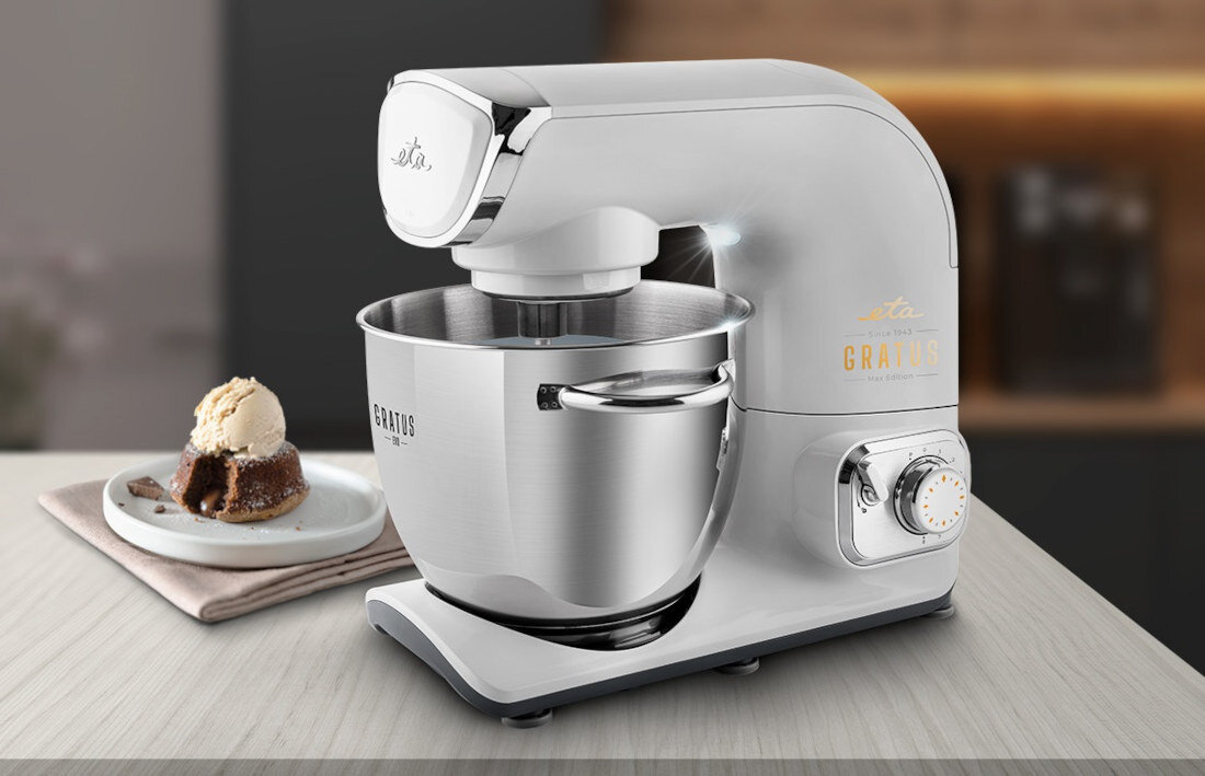 Na blacie kuchennym stoi srebrny robot kuchenny z misą, obok znajduje się talerz z ciastem i gałką lodów. Na robocie widoczne są napisy: \'eta\' i \'GRATUS\'.