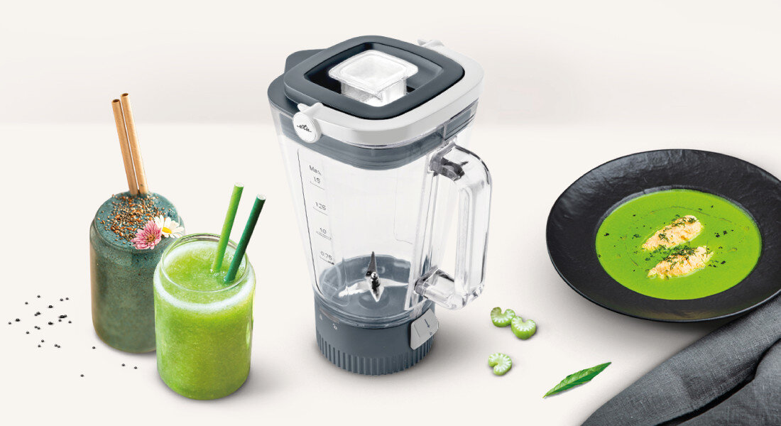 Na obrazie znajduje się przezroczysty blender kielichowy z miarką, obok którego stoją dwa zielone napoje w szklankach oraz talerz z zieloną zupą. Blender ma szary uchwyt i pokrywkę, a na miarce widoczne są oznaczenia w mililitrach.