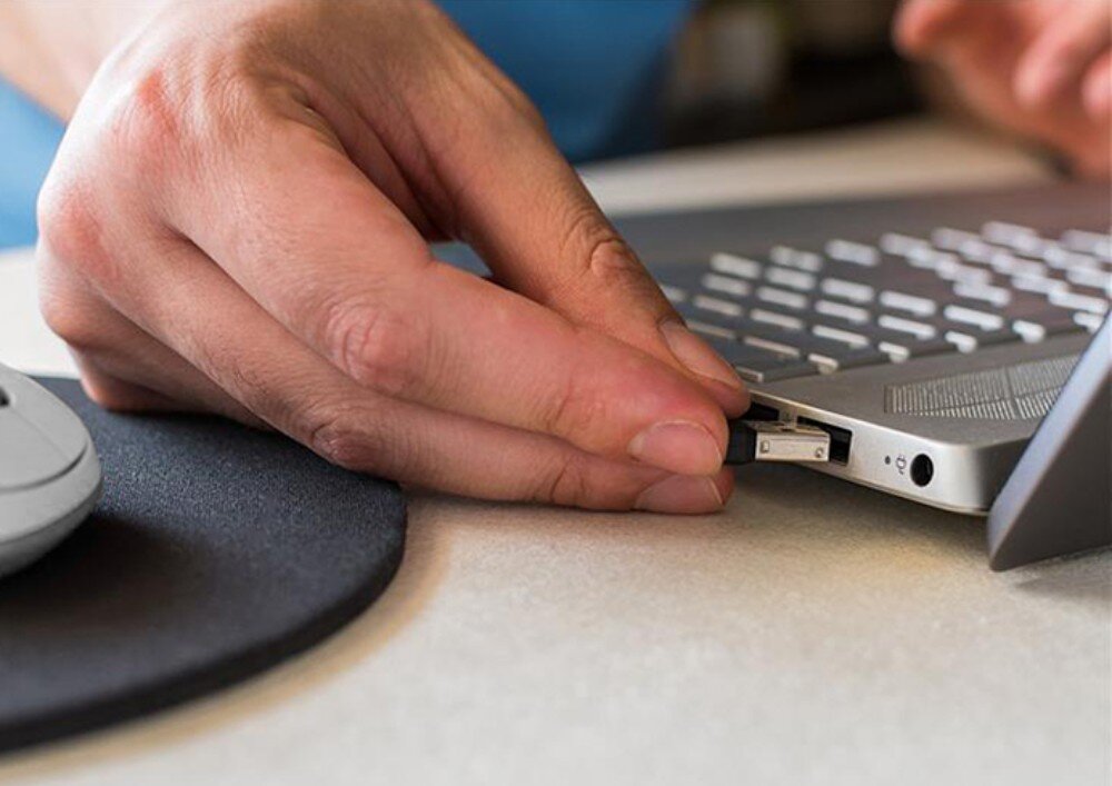 Osoba wkłada małe urządzenie USB do portu w laptopie. Obok znajduje się mysz komputerowa na podkładce.