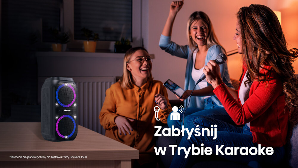 Na stole stoi czarny głośnik z dwoma podświetlanymi okręgami. Obok widoczne są trzy osoby, z których jedna trzyma mikrofon, a na obrazie znajduje się tekst: \'Zabłyśnij w Trybie Karaoke\'.