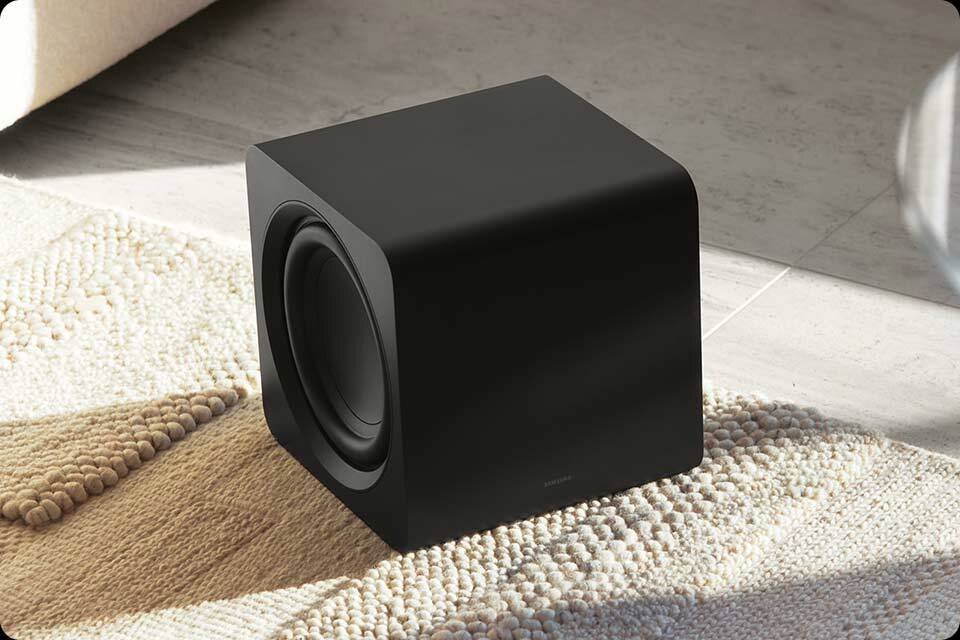Czarny subwoofer o prostokątnym kształcie stoi na jasnym dywanie z teksturą. Na dolnej krawędzi widoczne jest logo producenta.