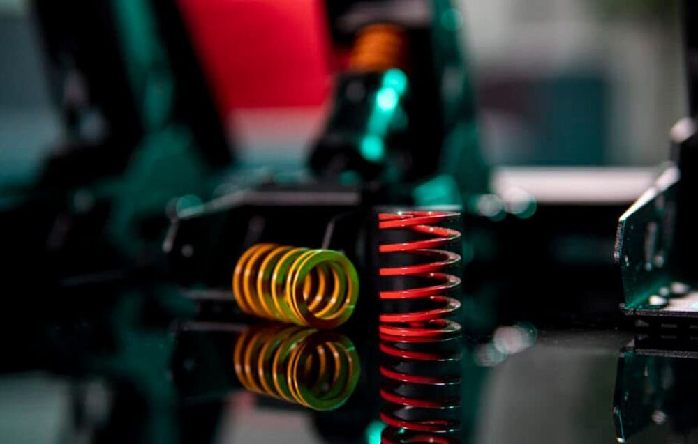 Na obrazie widoczne są dwie sprężyny, jedna w kolorze zielonym, druga czerwona, umieszczone na ciemnej powierzchni. W tle znajdują się elementy mechaniczne o metalicznym wyglądzie.