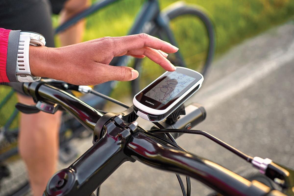 Osoba dotyka ekranu nawigacji rowerowej zamontowanej na kierownicy roweru. Na ekranie widoczna jest mapa, a na urządzeniu znajduje się logo \'Garmin\'.