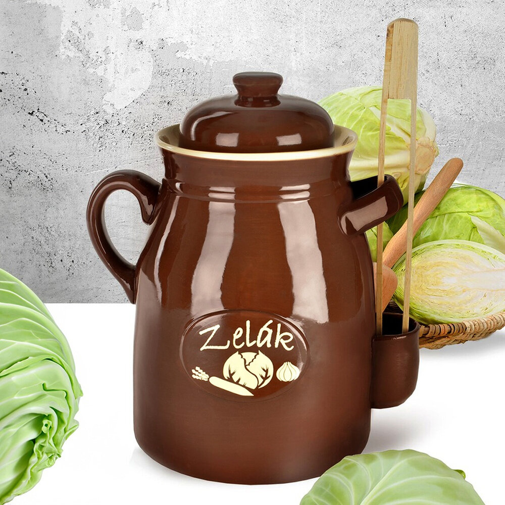 Brązowy ceramiczny pojemnik do kiszenia z pokrywką i szczypcami, z napisem \'Zelák\' oraz grafiką warzyw. W tle widoczne są główki kapusty na białym tle.