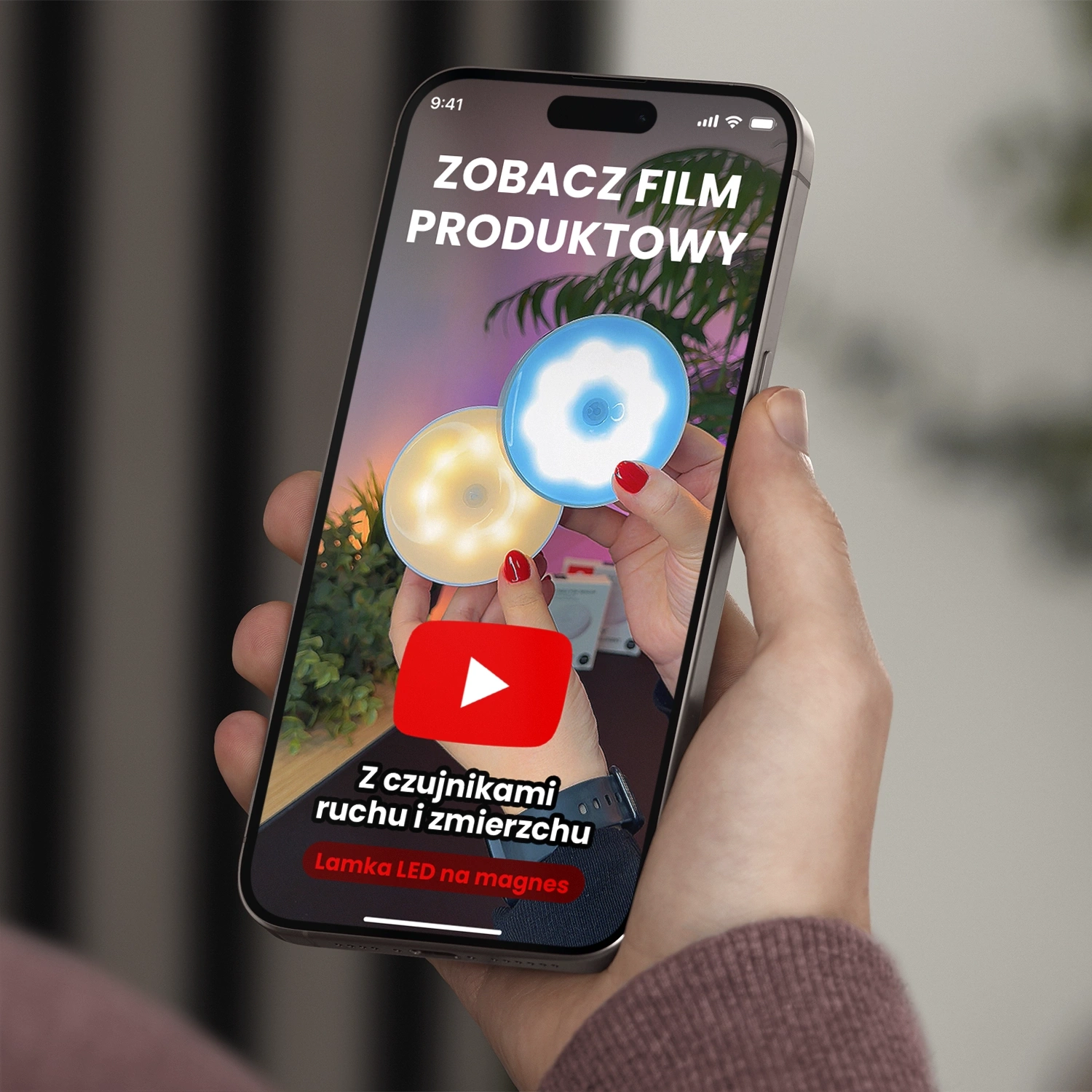 Osoba trzyma smartfon, na ekranie którego widnieje obraz dwóch okrągłych lamp LED w kolorach białym i niebieskim. Na ekranie znajduje się tekst: \'ZOBACZ FILM PRODUKTOWY\', \'Z czujnikami ruchu i zmierzchu\', \'Lampka LED na magnes\'.