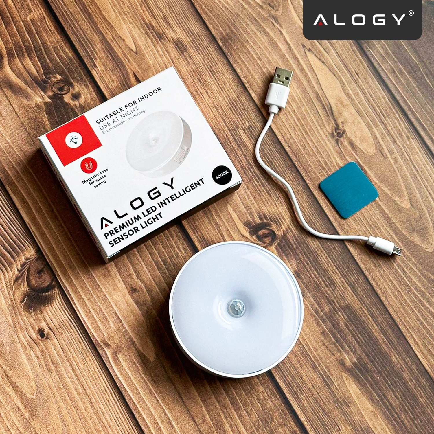 Na drewnianej powierzchni znajduje się okrągła lampka LED, pudełko z napisem \'Alogy Premium LED Intelligent Sensor Light\', biały kabel USB oraz niebieski kwadratowy element. Logo \'Alogy\' widoczne jest w rogu obrazu.