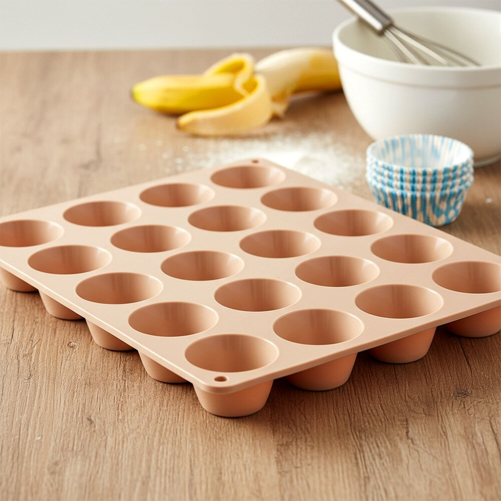 Na drewnianym blacie znajduje się beżowa silikonowa forma do muffinek z miejscem na 24 babeczki. W tle widoczne są banany, miska z trzepaczką oraz papierowe foremki do muffinek.