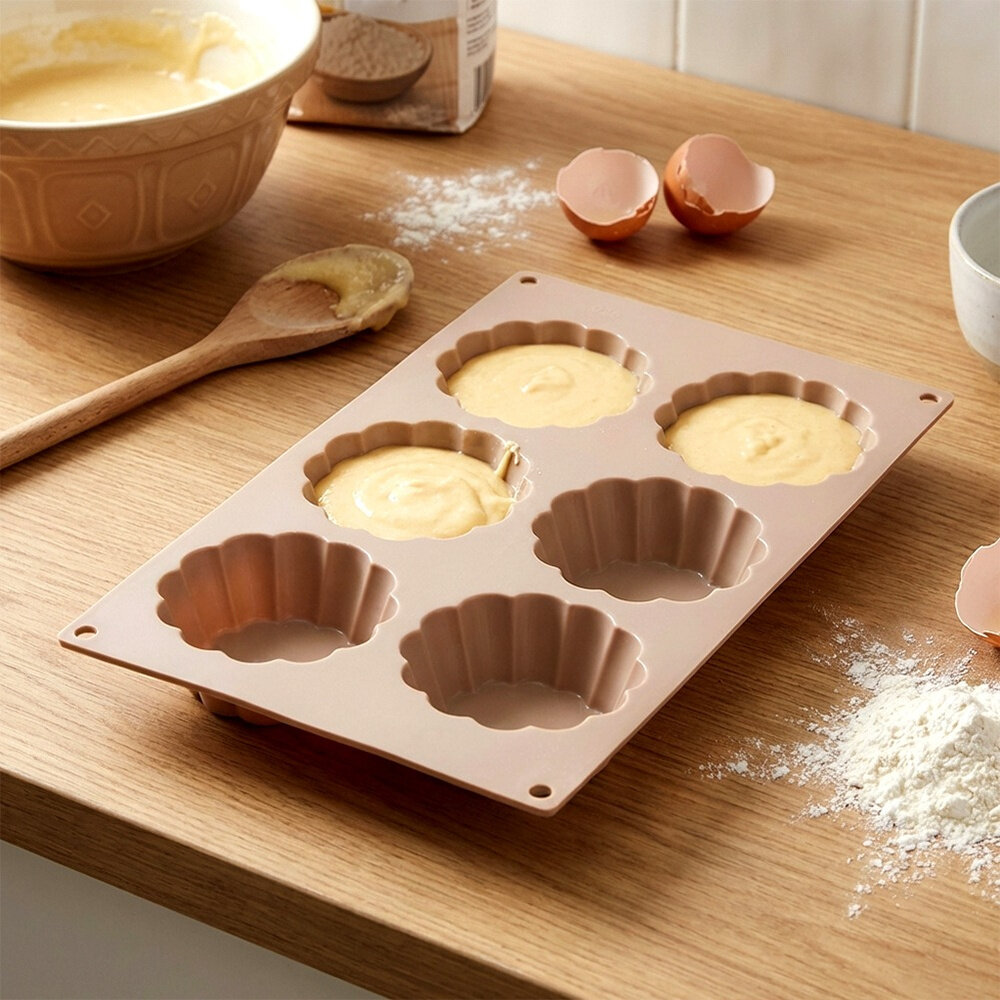 Na drewnianym blacie kuchennym znajduje się beżowa silikonowa forma do ciastek w kształcie kwiatków, wypełniona ciastem. Obok leży drewniana łyżka, miska z ciastem, rozbite skorupki jajek i rozsypana mąka.