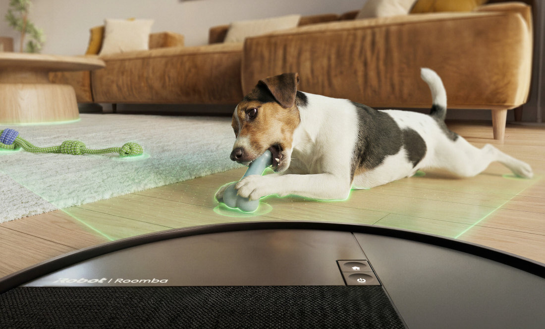 Na podłodze w salonie leży pies bawiący się zabawką, a w tle widać kanapę. Na pierwszym planie znajduje się fragment robota sprzątającego z widocznym logo \'iRobot Roomba\'.