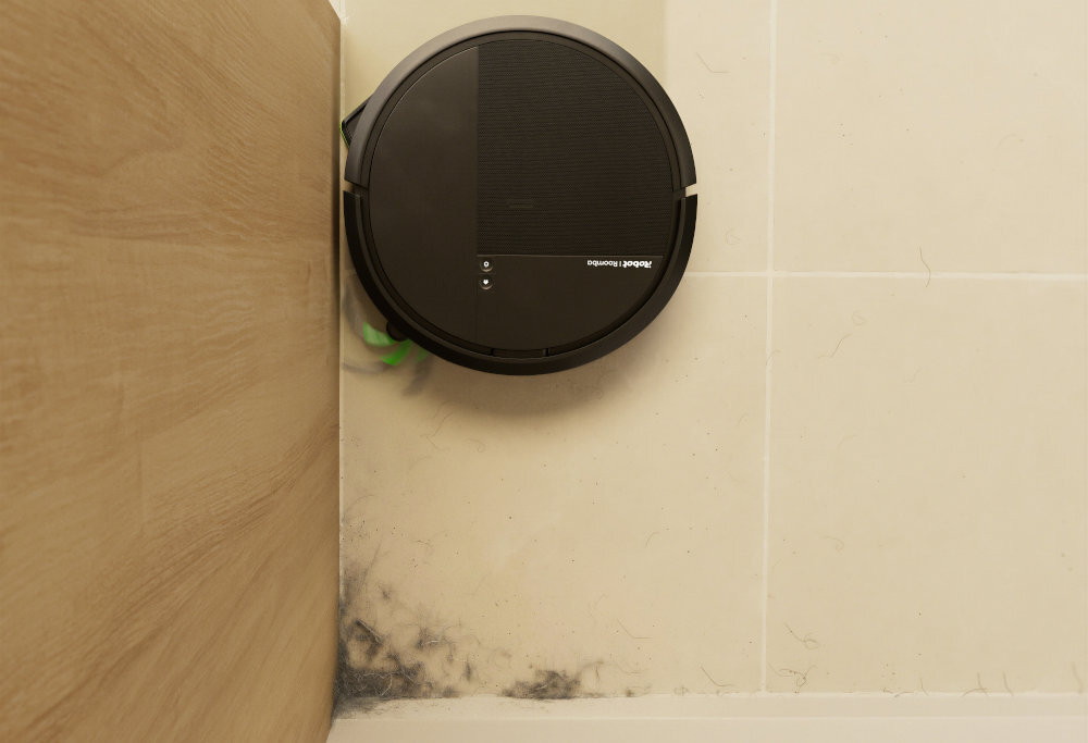 Czarny robot sprzątający znajduje się w rogu pomieszczenia na jasnej podłodze. Na urządzeniu widoczne jest logo \'iRobot Roomba\'.