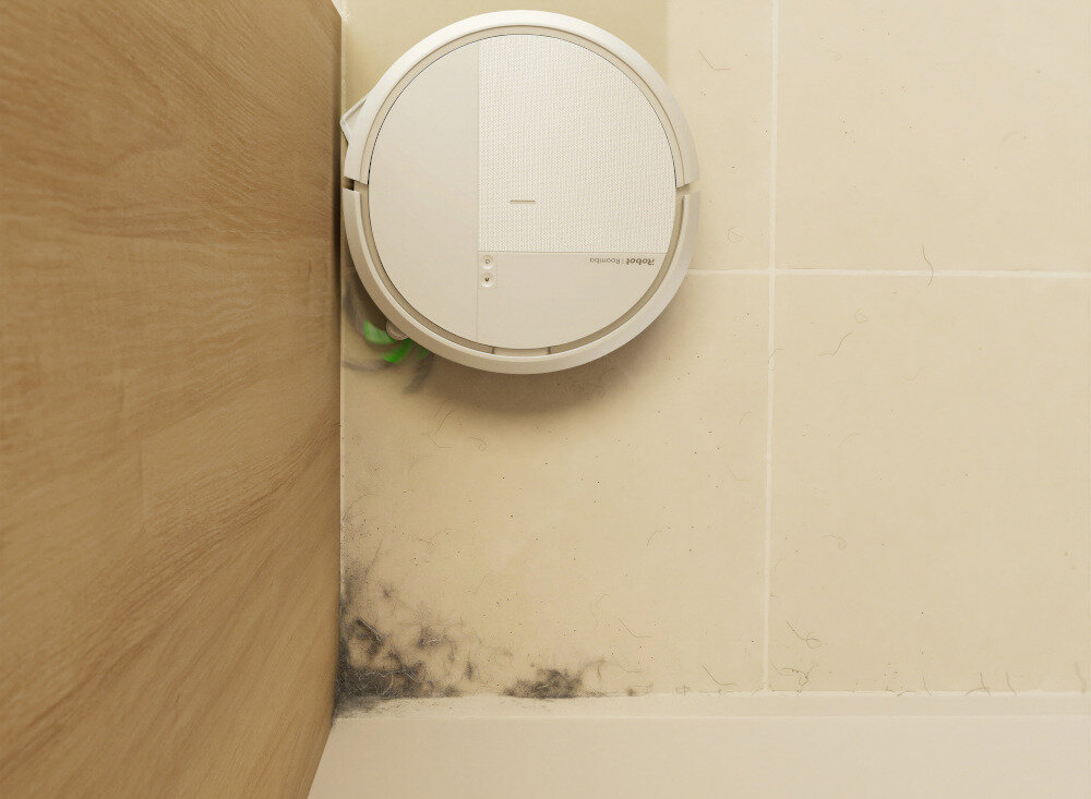 Na podłodze widać biały robot sprzątający z widocznym logo \'iRobot Roomba\'. Robot znajduje się w rogu pomieszczenia z jasnymi płytkami i drewnianą powierzchnią.