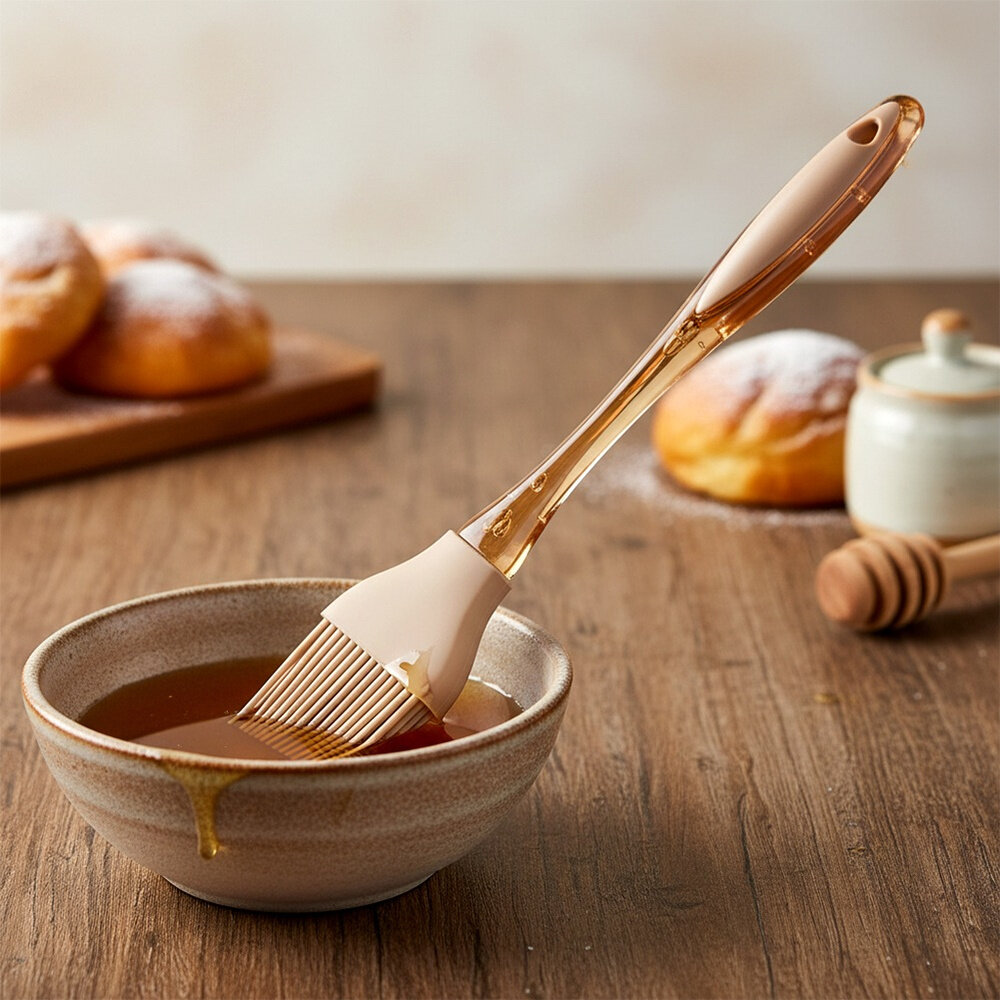 Na drewnianym stole znajduje się miska z płynem, w której zanurzony jest silikonowy pędzelek kuchenny. W tle widoczne są pączki i drewniany miodownik.