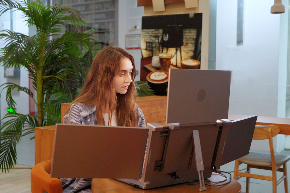 Osoba siedzi przy stole, przed nią znajduje się laptop z dwoma dodatkowymi monitorami. W tle widoczne są rośliny i dekoracje ścienne.