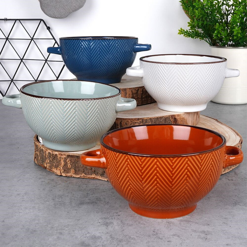 Na obrazie widoczne są cztery ceramiczne miseczki o pojemności 620 ml, każda w innym kolorze: niebieskim, białym, szarym i pomarańczowym, ustawione na drewnianych podstawkach. Miseczki mają delikatny wzór w jodełkę i dwa uchwyty po bokach.
