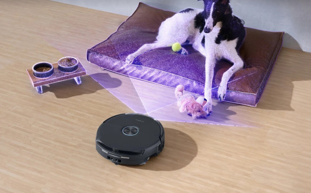 Na podłodze znajduje się czarny robot sprzątający, a obok leży pies na fioletowej poduszce z zabawką i miskami. Robot emituje niebieskie światło na podłogę.