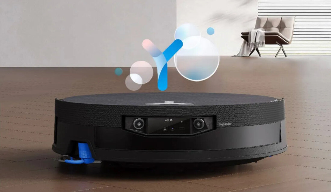 Na podłodze znajduje się czarny robot sprzątający z widocznymi kamerami i napisem \'AIVI 3D\' oraz \'FocusJet\'. W tle widać nowoczesne wnętrze z krzesłem i dekoracyjnymi elementami.