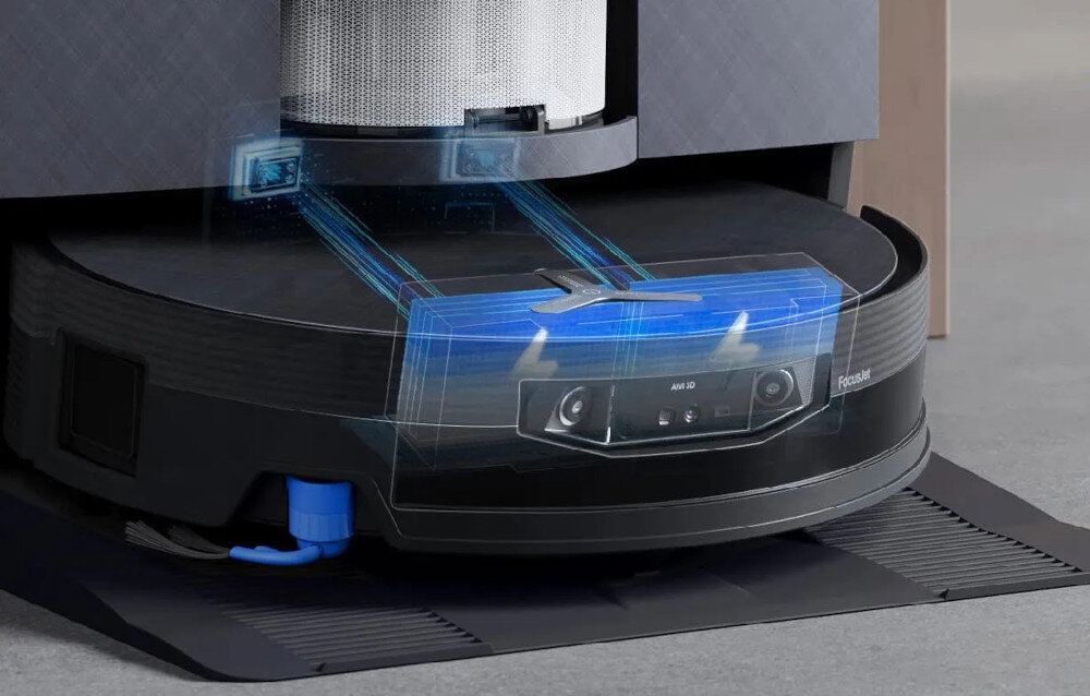 Na obrazie widoczny jest robot sprzątający z elementami technologicznymi, takimi jak czujniki i filtry. W tle znajduje się stacja dokująca z metalowym filtrem.