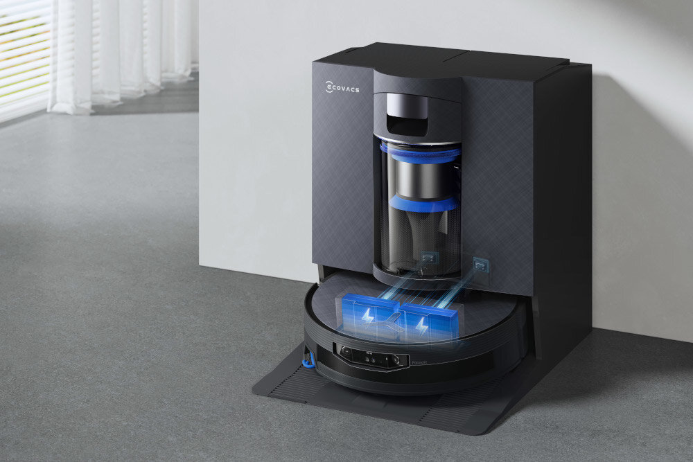 Na obrazie widoczny jest czarny robot sprzątający w stacji dokującej z logo \'ECOVACS\'. W tle znajduje się jasna podłoga i zasłony.