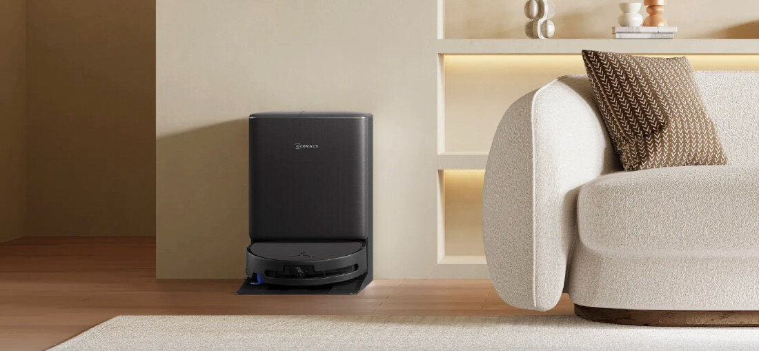 Na obrazie widać robot sprzątający w stacji dokującej z logo \'ECOVACS\', ustawiony na drewnianej podłodze obok jasnej sofy z poduszką. W tle znajduje się półka z dekoracjami.