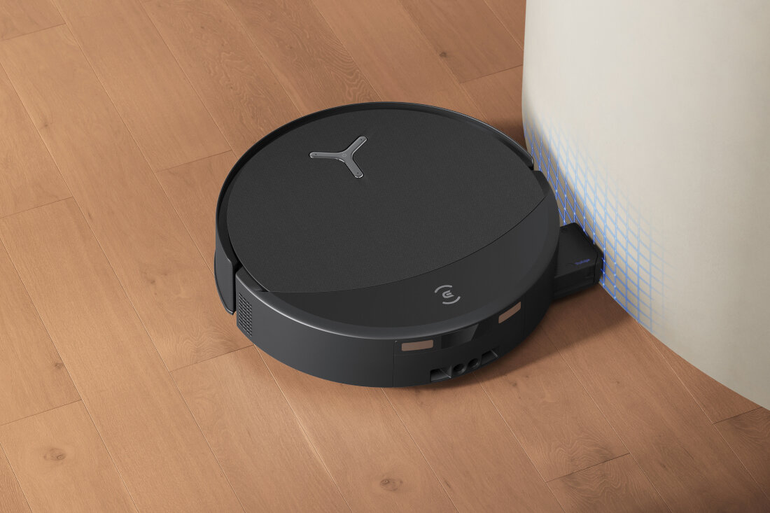 Czarny robot sprzątający o okrągłym kształcie porusza się po drewnianej podłodze wzdłuż jasnej ściany. Na górnej powierzchni robota widoczne jest logo w kształcie litery \'Y\'.