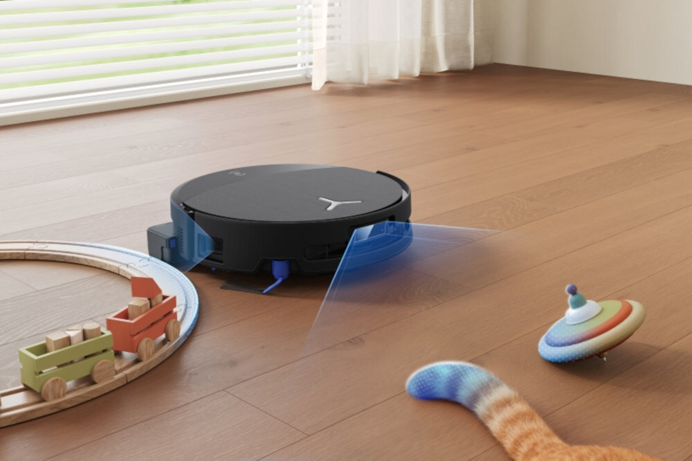 Na drewnianej podłodze znajduje się okrągły robot sprzątający z widocznym niebieskim światłem. Obok robota leżą zabawki: drewniany pociąg na torach i kolorowy bączek.