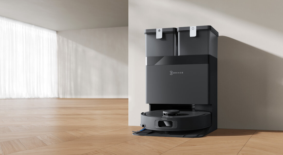 Na drewnianej podłodze stoi czarny robot sprzątający z logo \'ECOVACS\' na stacji dokującej. W tle widoczne są jasne zasłony i ściana.