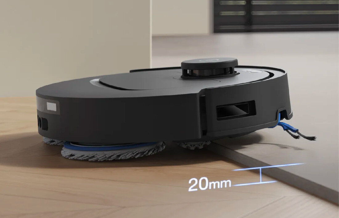 Czarny robot sprzątający pokonuje próg o wysokości 20 mm na drewnianej podłodze. Widoczne są szczotki czyszczące oraz napis \'20 mm\'.