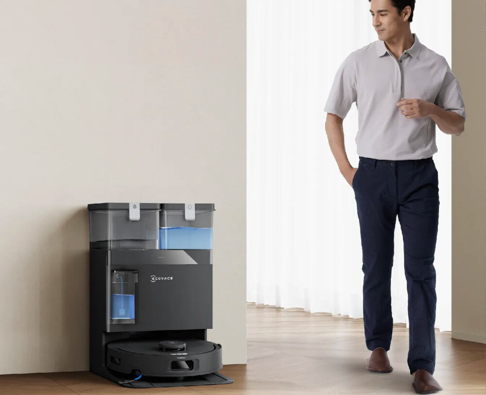 Na obrazie widoczny jest robot sprzątający z czarną stacją dokującą marki ECOVACS. Obok stoi osoba ubrana w jasną koszulę i ciemne spodnie.