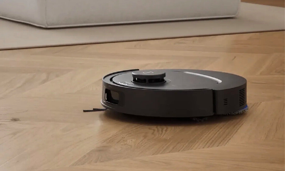 Na drewnianej podłodze znajduje się okrągły, czarny robot sprzątający. W tle widoczna jest jasna sofa.
