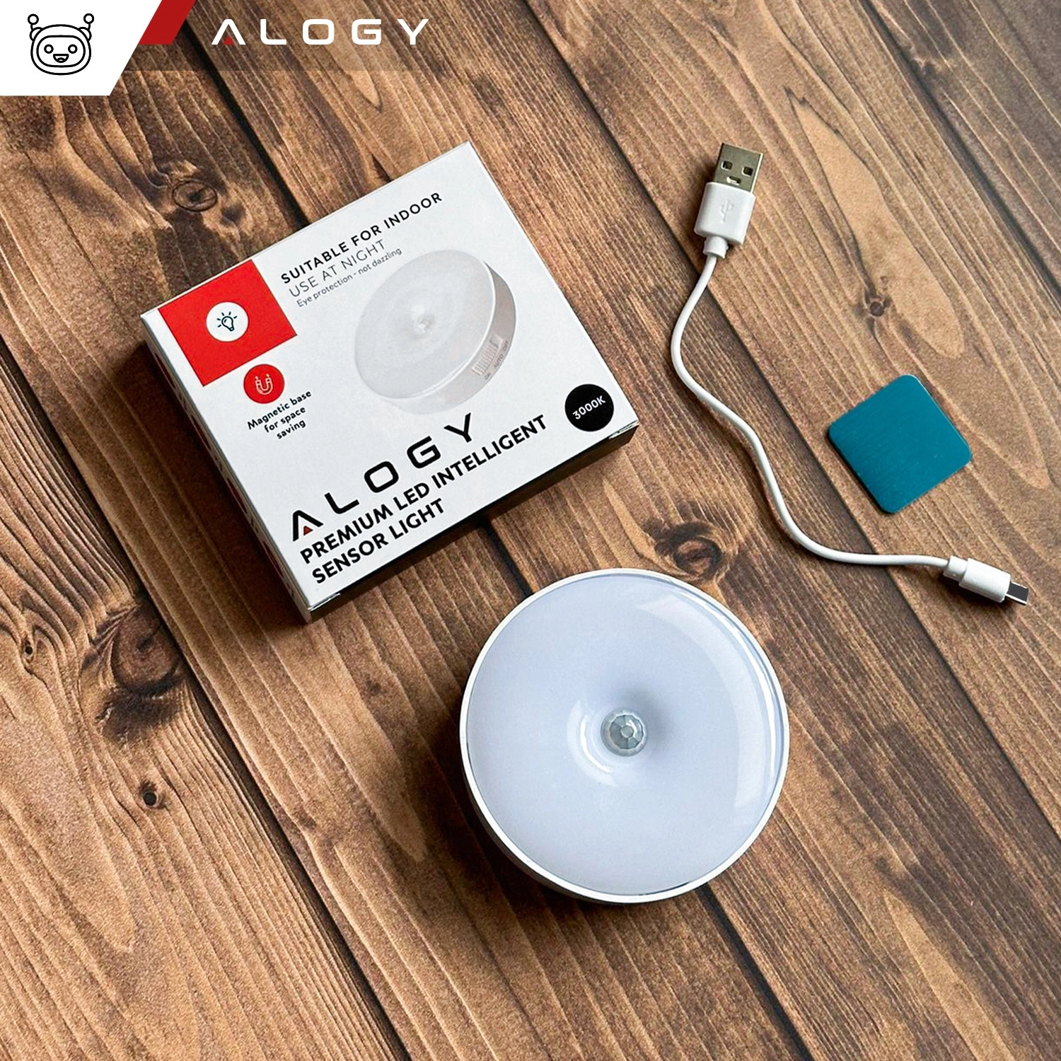 Na drewnianej powierzchni znajduje się okrągła lampka LED, pudełko z napisem \'Alogy Premium LED Intelligent Sensor Light\', biały kabel USB oraz niebieski kwadratowy element. Na pudełku widnieje napis \'Suitable for indoor use at night\' oraz logo Alogy.