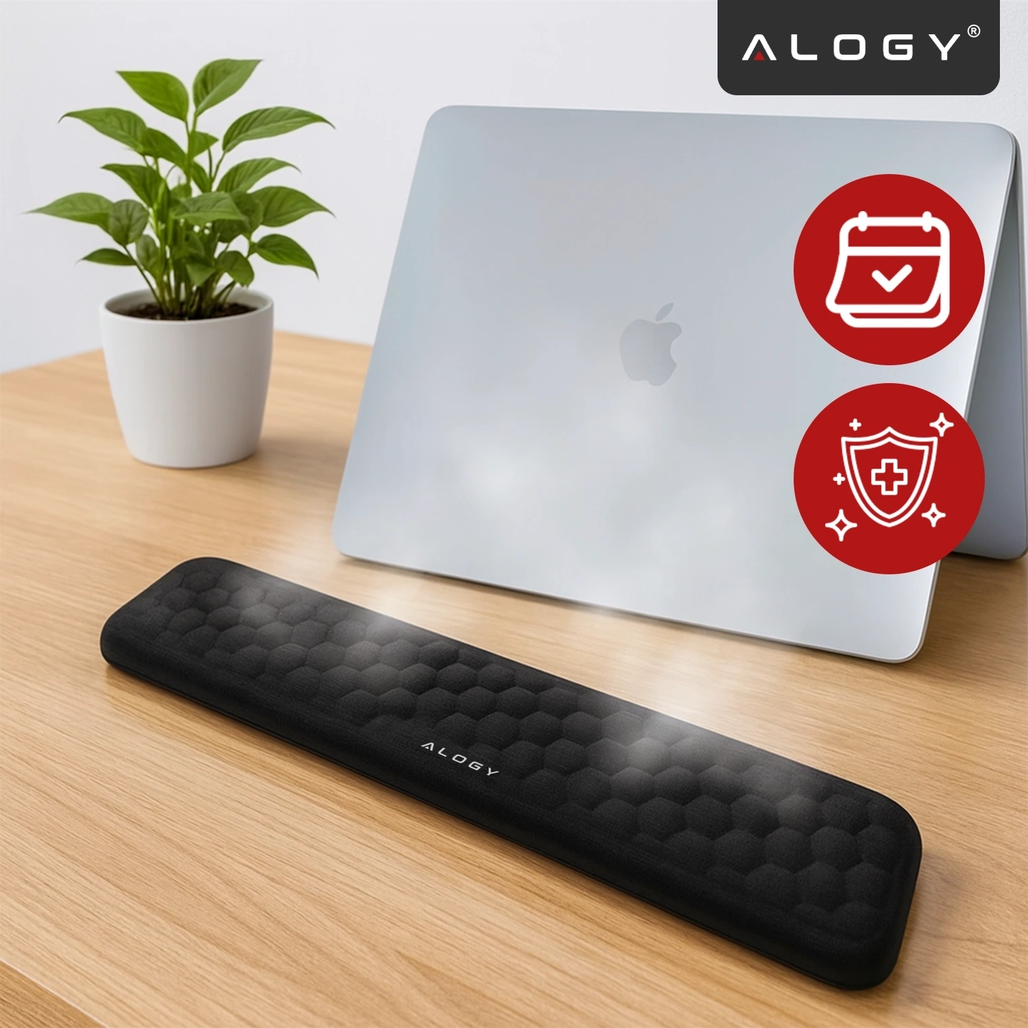 Na drewnianym biurku znajduje się czarna podkładka pod nadgarstki z napisem \'ALOGY\', obok srebrnego laptopa z logo jabłka. W tle widoczna jest biała doniczka z zieloną rośliną oraz dwa czerwone symbole: kalendarz z zaznaczeniem i tarcza z krzyżem.