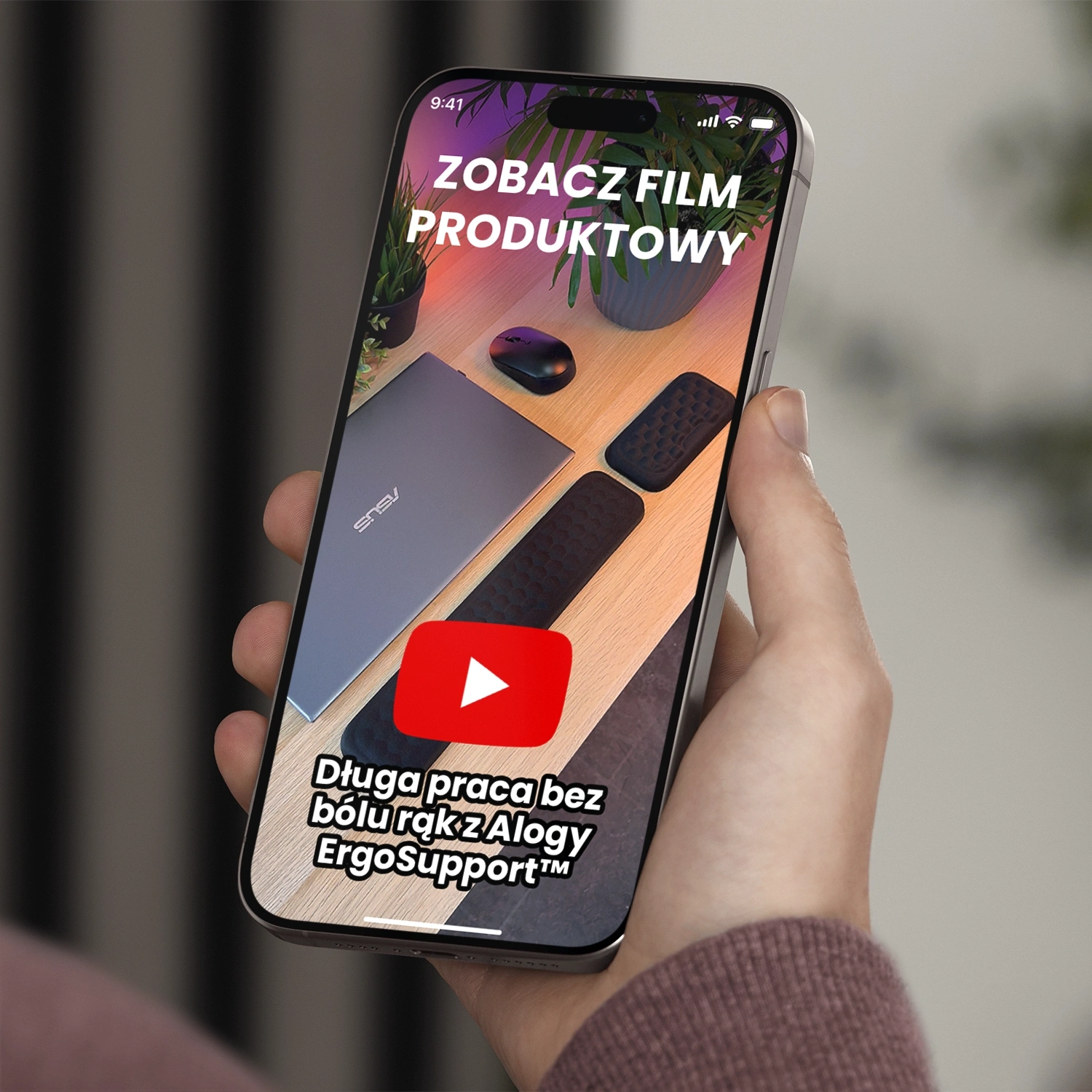 Osoba trzyma telefon, na ekranie którego widać zdjęcie biurka z laptopem, myszką i podkładkami pod nadgarstki. Na ekranie widoczny jest tekst: \'ZOBACZ FILM PRODUKTOWY\' oraz \'Długa praca bez bólu rąk z Alogy ErgoSupport™\'.