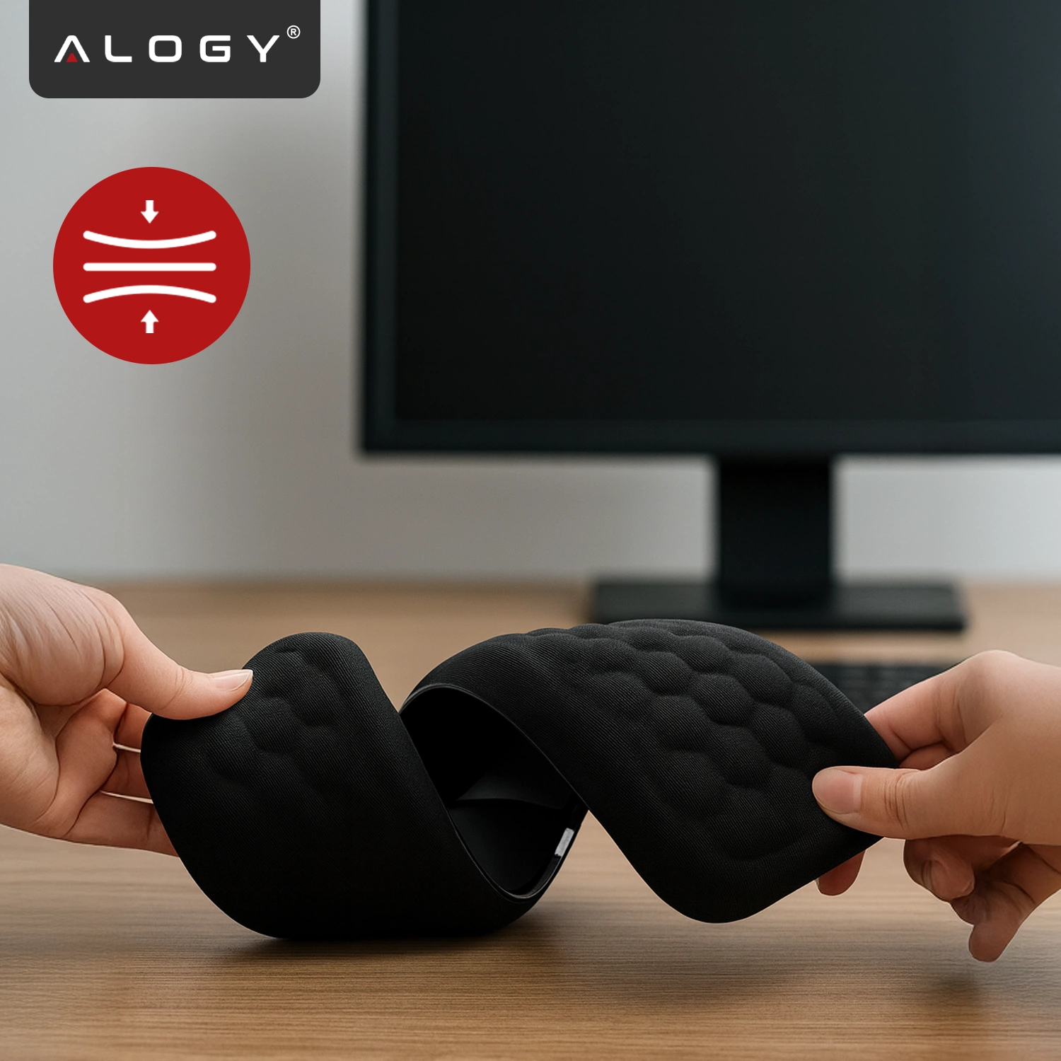 Na drewnianym biurku znajduje się czarna podkładka z pianki memory, trzymana przez dwie ręce. W tle widoczny jest monitor, a w lewym górnym rogu logo \'ALOGY\'.