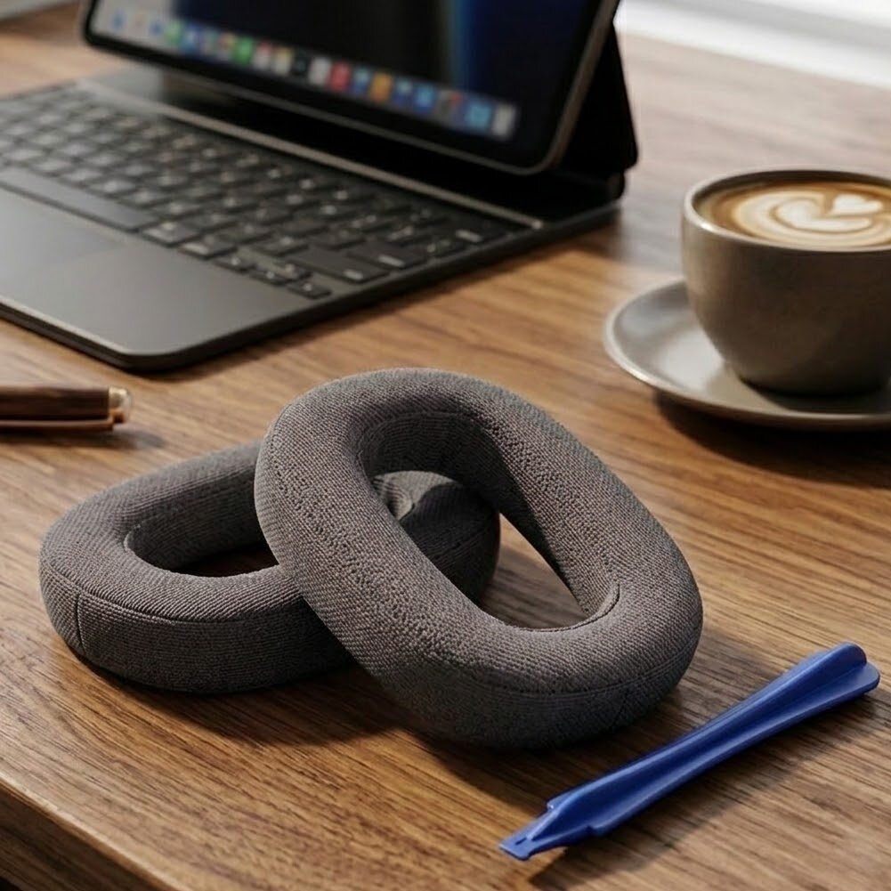 Na drewnianym stole leżą szare nauszniki oraz niebieskie narzędzie. W tle znajduje się otwarty laptop i filiżanka kawy z mleczną pianką.