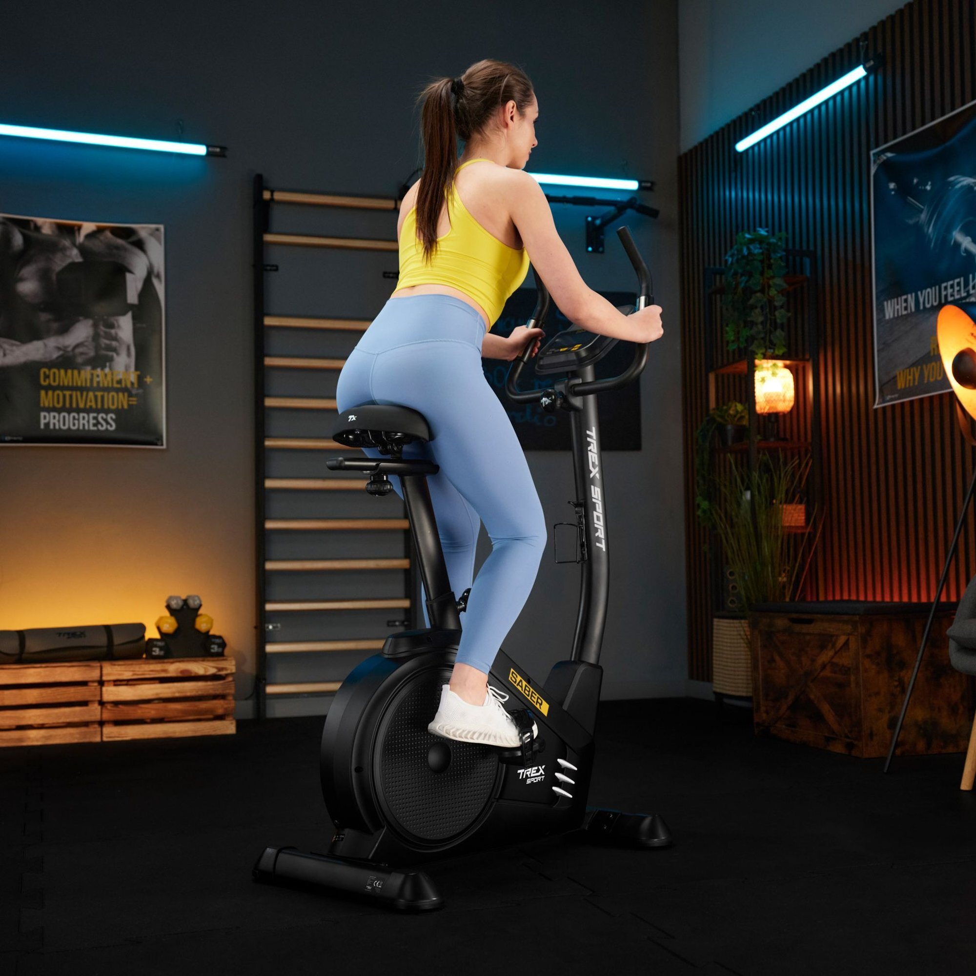 Osoba ćwiczy na czarnym rowerze stacjonarnym elektryczno-magnetycznym SABER TX-750EB w pomieszczeniu z plakatami i sprzętem do ćwiczeń. Na rowerze widoczne są napisy \'SABER\' i \'TREX SPORT\'.