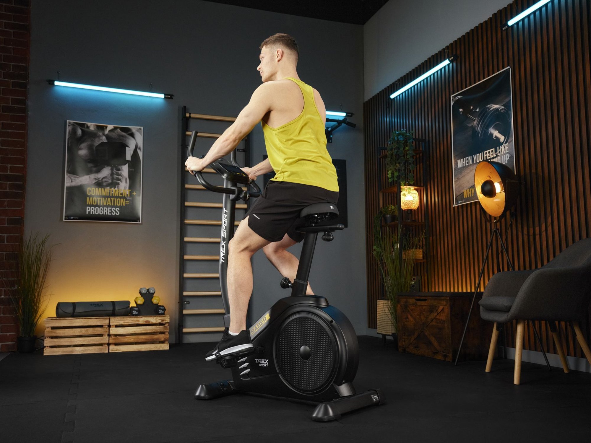 Osoba ćwiczy na czarnym rowerze stacjonarnym elektryczno-magnetycznym SABER TX-750EB w pomieszczeniu z plakatami na ścianach i sprzętem do ćwiczeń. Na rowerze widoczne jest logo \'media expert\'.